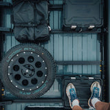 Top-down view of a Front Runner Toyota HiAce LWB (2019-Current) Slimpro Roof Rack Kit with a spare tire mounted on the left, a black bag hanging at the top left, and a black hard case at the top right. A person's legs and shoes are visible at the bottom right standing on a black storage box.