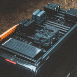A Front Runner Toyota HiAce LWB (2019-Current) Slimpro Roof Rack Kit loaded with camping gear, including hard-sided storage cases, a duffel bag, and a solar panel, shown from an elevated rear angle on a vehicle outdoors.