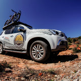 A white Isuzu pickup truck parked on a rocky desert road under a clear blue sky, with a Front Runner Series Isuzu MU-X (2017-2020) Slimline II Roof Rack Kit and roof-mounted bike rack carrying two bicycles. The side of the truck has a large circular logo reading 'BEYOND WILD' and 'ISUZU ADVANTER' with a website printed on the door.