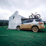 A muddy white SUV parked on green grass in front of a white stucco building, with a mountain bike strapped to its Front Runner Series Isuzu MU-X (2017-2020) Slimline II Roof Rack Kit. The vehicle has graphic decals on the side and rear.
