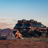 A woman sits inside a gray pop-up camping tent pitched beside two dark SUVs with Front Runner Series Isuzu MU-X (2021-Current) Slimline II Roof Rack Kit and camping gear, at an outdoor campsite during late afternoon under a blue sky.