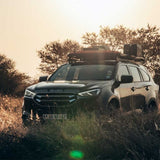A black Isuzu SUV parked outdoors in a grassy field at sunset, with trees in the background and Front Runner Series Isuzu MU-X (2021-Current) Slimline II Roof Rack Kit with mounted cargo gear visible.