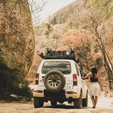 A woman walks beside a white off-road SUV with a Front Runner Suzuki Jimny (1998-2018) Slimline II Roof Rack Kit and spare tire, parked on a dirt road in a rocky, tree-filled outdoor setting.