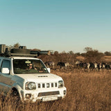 A white safari vehicle with a Front Runner Suzuki Jimny (1998-2018) Slimline II Roof Rack Kit and spare tire is parked in a dry grass field, with zebras visible in the background under a clear blue sky.