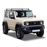 A beige Suzuki Jimny SUV with a black Front Runner Suzuki Jimny (2018-Current) Load Bar Kit mounted on the roof, black front and side bumpers, and black wheels, shown from a three-quarter front angle on a white background.