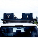 A black Front Runner Toyota Land Cruiser 300 Slimline II Roof Rack Kit mounted on the roof of a white vehicle, holding two dark hard-sided storage boxes under an overcast sky.