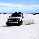 A white Toyota off-road SUV drives across bright white sand dunes, kicking up a large cloud of dust under a blue sky. Front Runner Toyota Land Cruiser 300 Slimline II Roof Rack Kit is mounted on the roof