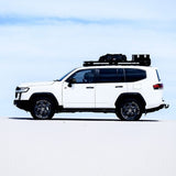 A white SUV shown in side profile on a bright white surface under a pale blue sky, with a Front Runner Toyota Land Cruiser 300 Slimline II Roof Rack Kit carrying black gear.