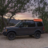 A dark gray SUV parked outdoors on a gravel surface under trees at sunset, with Front Runner Series Recovery Device & Gear Holding Side Brackets mounted on top.
