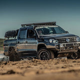 A dark gray lifted Volkswagen pickup truck with a roof rack and front bumper kit driving through mud on a sandy off-road trail. The truck is equipped with Front Runner Series Recovery Device & Gear Holding Side Brackets