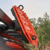 Bright orange roof rack mounting accessories with black Front Runner Series Recovery Device & Gear Holding Side Brackets, shown attached to a vehicle roof rack outdoors with trees in the background.