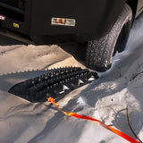 A black MAXTRAX XTREME next to a black vehicle tire, secured with an orange strap.