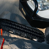 A close-up of a black MAXTRAX XTREME tire tread with a textured, knobby tread pattern, positioned next to a white vehicle on a sandy surface with a gray tarp.