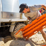 A man in a gray t-shirt and black cap pushes an orange MAXTRAX XTREME  recovery board loaded with sand over a rugged off-road tire of a white truck in a sandy outdoor setting.
