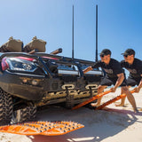 Two men in black shirts and caps are manually rolling bright orange MAXTRAX XTREME tracks with metal spikes up a sandy incline next to a black off-road vehicle on a desert beach under a clear blue sky.