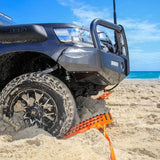 An orange MAXTRAX XTREME traction mat with a black handle is deployed from under a dark gray off-road vehicle on a sandy beach with the ocean and blue sky in the background.