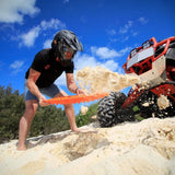 A man wearing a black helmet and black t-shirt uses a bright orange MAXTRAX Mini and Jaxbase Combo to push sand aside next to a red and black off-road vehicle on a sandy beach under a blue sky.