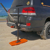 An orange MAXTRAX Mini and Jaxbase Combo is use as a solid base on the sand in front of a black Toyota Land Cruiser SUV to jack up the vehicle on a beach.