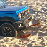 A blue off-road vehicle with a black front bumper is driving through sandy terrain, with two MAXTRAX Mini and Jaxbase Combo embedded in the sand nearby.