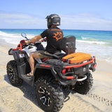 A person rides a black and white Can-Am ATV on a sandy beach with the ocean in the background. The ATV has orange accents and a bright orange MAXTRAX Mini and Jaxbase Combo on the back.