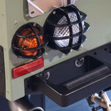 Close-up of a vehicle rear light assembly with two black protective cage-mounted lights, one amber and one clear/white, plus a red reflector and a black bumper.