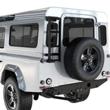 A white off-road vehicle shown from the rear three-quarter angle, featuring a Front Runner Land Rover Defender 90/110 (1983-2016) Ladder mounted on the side and a spare tire mounted on the rear door.