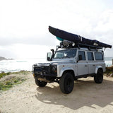 Land Rover fitted with the Front Runner Land Rover Defender 110 (1983-2016) Slimline II Roof Rack Kit  with a kayak on top parked near a beach