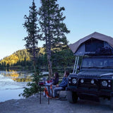Two people sitting at a table by a lake with a rooftop tent on a vehicle.
