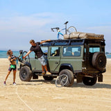 Two people with a vintage truck fitted with the Front Runner Land Rover Defender 110 (1983-2016) Slimline II Roof Rack Kit on a beach