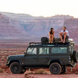 Two people sitting on the roof of a Front Runner Land Rover Defender 110 (1983-2016) Slimline II Roof Rack Kit  in a desert landscape with mountains in the background.