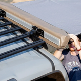 A tan roof-mounted awning with black roof racks and Front Runner Series Slimsport Side Mount Accessory Bracket / Small, installed on a light-colored vehicle, with a man adjusting the fabric in the background.