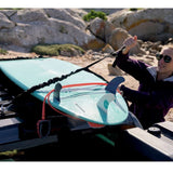 A woman wearing sunglasses and a purple jacket secures a light blue surfboard strapped to the back of a vehicle with a Front Runner Stratchits, set against a rocky beach background.