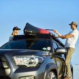 A man and a woman are loading a black and red kayak onto a Front Runner roof rack of a dark gray SUV on a beach under a clear blue sky.