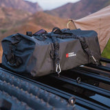A black Front Runner duffel bag with a white and red logo strapped to a roof rack with a Front Runner Stratchits tie down strap, resting on a black vehicle roof rack with a mountainous outdoor background.