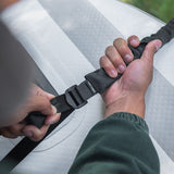 A close-up of a person's hands adjusting a black Front Runner Stratchits tie down strap with a plastic buckle over a white textured surface.