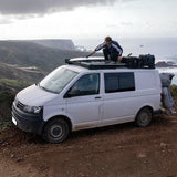 A white camper van parked on a dirt road with a person crouching on top of the Front Runner Volkswagen T5 Transporter SWB (2003-2015) Slimline II Roof Rack Kit fitted to the van, with a rocky coastal landscape and ocean in the background.