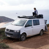 A white Volkswagen camper van parked on a dirt cliffside road with the ocean and coastline in the background. A person is sitting on the Front Runner Volkswagen T5 Transporter SWB (2003-2015) Slimline II Roof Rack above the vehicle.
