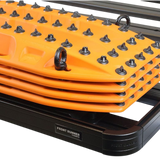 A stack of bright orange Maxtrax with black spikes and metal D-rings, shown resting on a black Roof Rack, using the Front Runner Series Recovery Device Mounting Kit. A label on the frame reads 'FRONT RUNNER'.