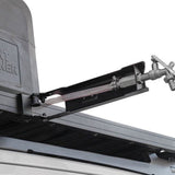 Close-up of a black metal Front Runner Tap Extension Bracket and hose assembly attached to a vehicle, with part of a gray container visible on the left against a white background.