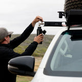 A man in a black jacket and gray cap fills a bottle using the Front Runner Tap Extension Bracket attached to the roof rack of a white vehicle outdoors.