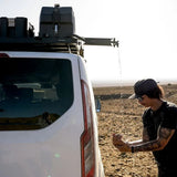 A man in a black cap and sunglasses washes his face with water dripping from a roof rack-mounted Front Runner Tap Extension Bracket and hose on a white vehicle in an open desert landscape.