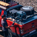 A man in a black cap and dark shirt is filling a black water bottle from a Front Runner 42L water tank and Tap Extension Bracket a black roof rack system mounted on a red Ford Ranger pickup truck. The truck bed is loaded with a spare tire, a green water container, and black storage cases, set against a rocky outdoor background.