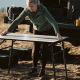 A woman outdoors at sunset adjusts a Front Runner Under Rack Table with a dark gray top and black metal legs, with camping gear and a vehicle in the background.