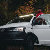 A white Volkswagen van parked outdoors in a wooded area with autumn foliage, with a person in a red jacket standing beside the open driver's side door. A bright roof-mounted light bar is illuminated above the vehicle on a Front Runner Front Runner Volkswagen T5/T6/T6.1 Transporter SWB (2003-2024) Slimsport Roof Rack Kit.