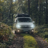 A white Volkswagen camper van with a Front Runner Front Runner Volkswagen T5/T6/T6.1 Transporter SWB (2003-2024) Slimsport Roof Rack Kit is driving on a dirt path through a dense forest, with its headlights on.