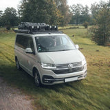 A silver Volkswagen camper van with a Front Runner Front Runner Volkswagen T5/T6/T6.1 Transporter SWB (2003-2024) Slimsport Roof Rack Kit and camping setup, driving on a grassy outdoor path with trees in the background.