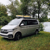 A silver Volkswagen camper van parked on grass beside a lake, with trees in the background and a yellow and white kayak strapped to the Front Runner Front Runner Volkswagen T5/T6/T6.1 Transporter SWB (2003-2024) Slimsport Roof Rack Kit.