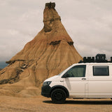 A white camper van with black wheels and Front Runner Front Runner Volkswagen T5/T6/T6.1 Transporter SWB (2003-2024) Slimsport Roof Rack Kit parked on a dirt road in front of a large sandstone badlands rock formation under a cloudy sky.