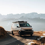 A beige Volkswagen Transporter camper van with a Front Runner Volkswagen T6/T6.1 Caravelle Transporter SWB (2015-Current) Slimline II Roof Rack Kit is parked on a dirt mountain road with snowy peaks in the background.