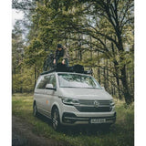 A white Volkswagen camper van parked on a grassy forest road, with a person standing on top of the vehicle and packing gear onto the Front Runner Volkswagen T6/T6.1 Caravelle Transporter SWB (2015-Current) Slimline II Roof Rack Kit. Trees and greenery fill the background.